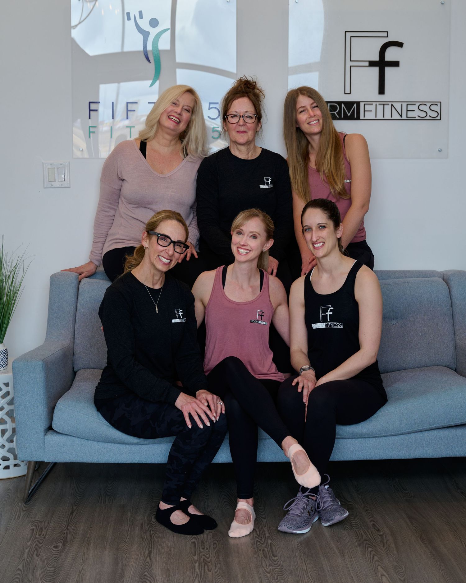 A group of fitness instructors pose together on a gray couch in a bright studio with studio logo on the wall.