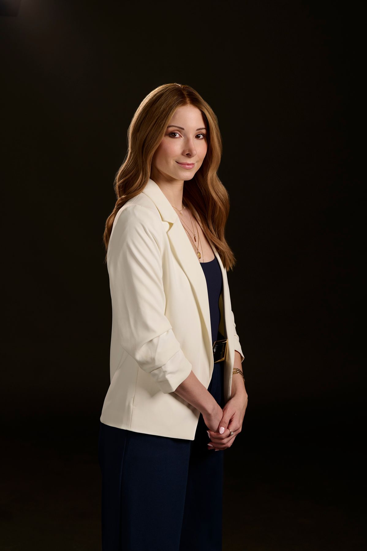 Professional studio portrait of someone wearing a white blazer and navy dress against a dark background.