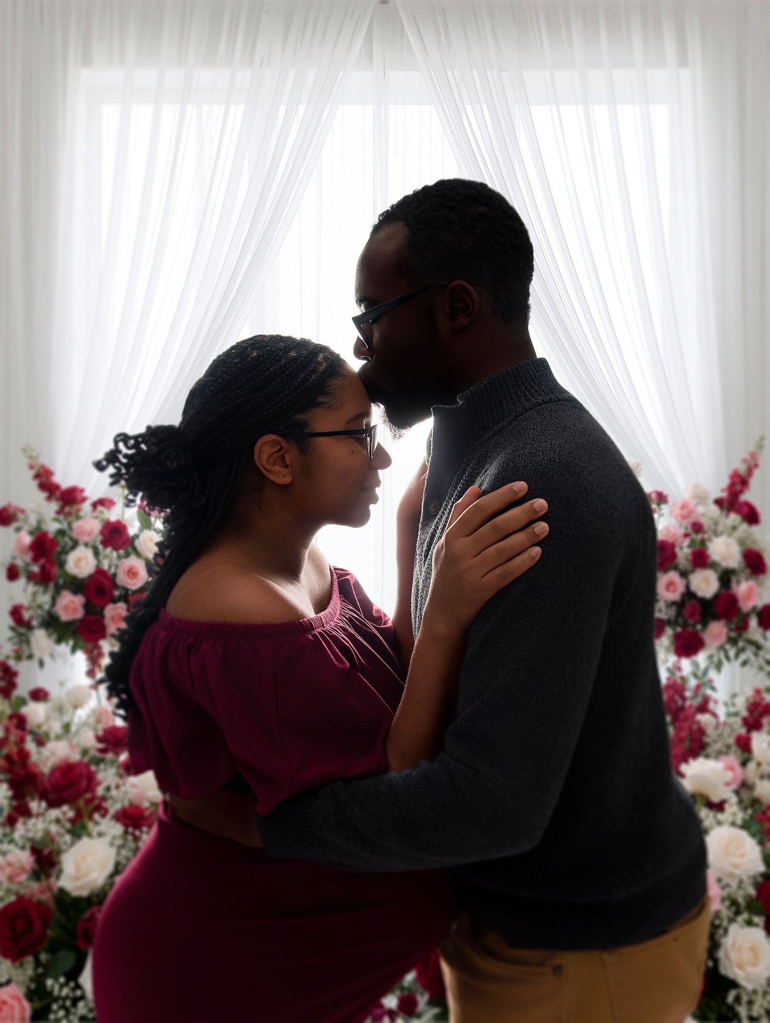 Couple sharing a romantic moment surrounded by pink and white floral arrangements near a sheer curtained window.