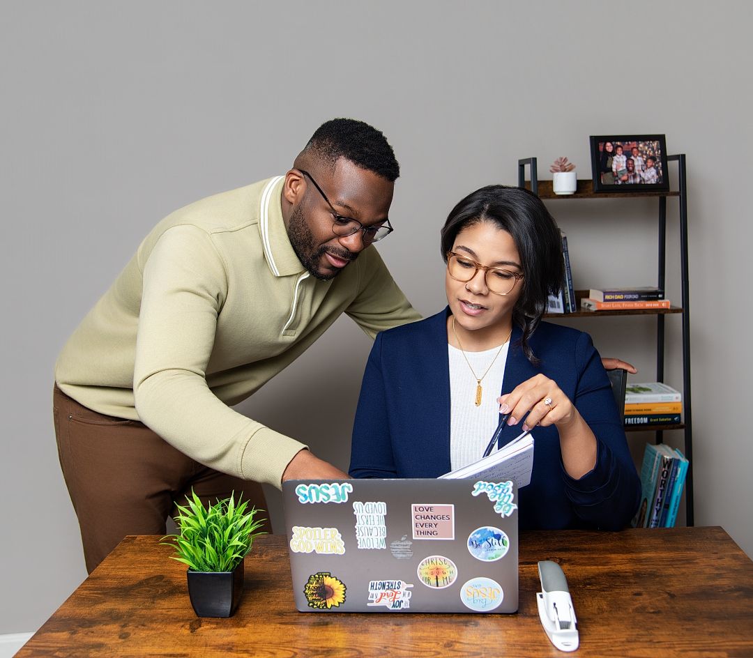 Two professionals collaborating on a laptop with stickers at a wooden desk in a modern office setting.
