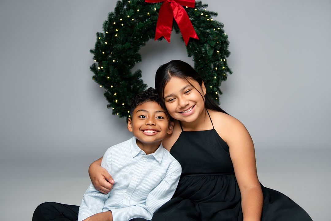 Two people pose together in front of a festive green Christmas wreath with a red bow against a gray backdrop.