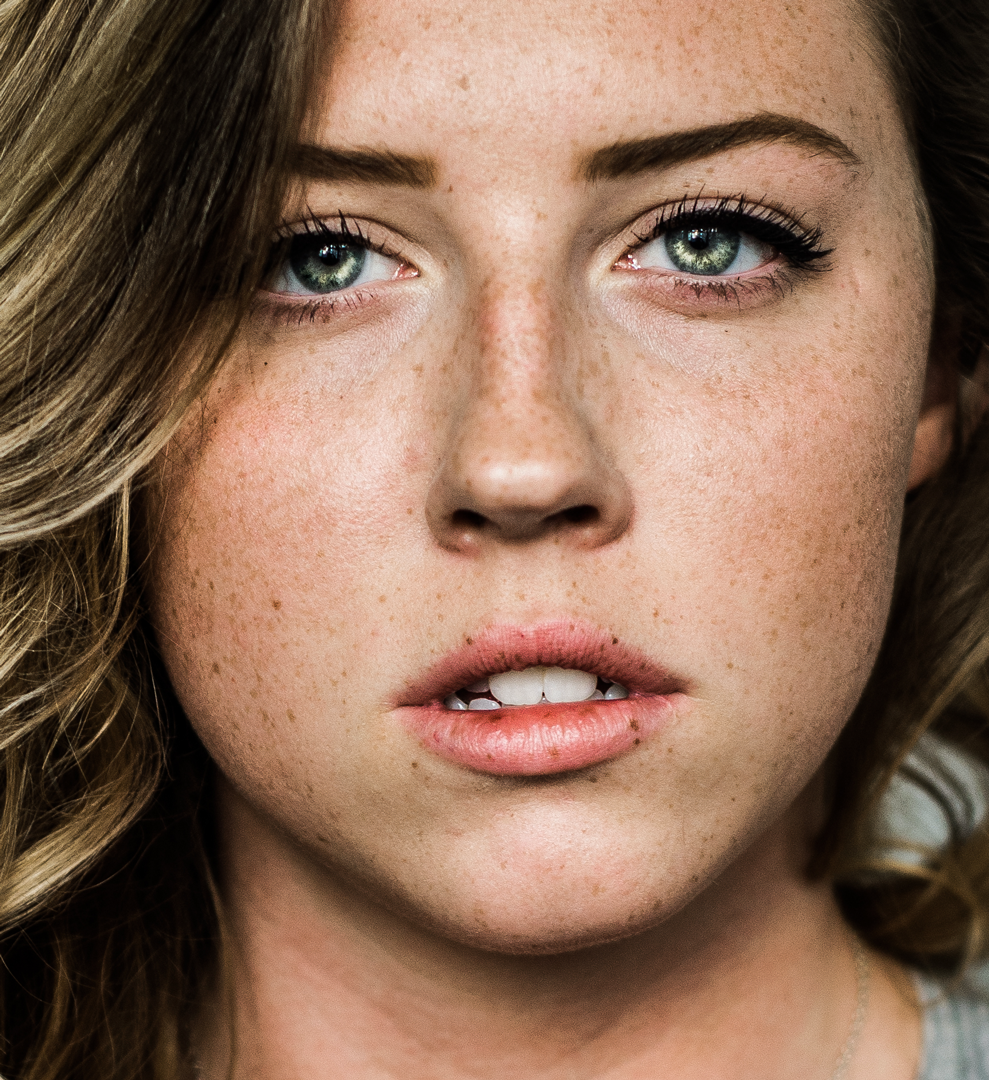 Close-up portrait of a young woman with green eyes, freckles, and blonde hair gazing directly at the camera.