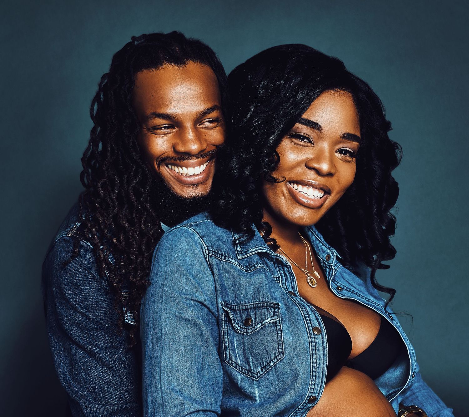 Couple in denim jackets sharing joyful laughter against dark background.