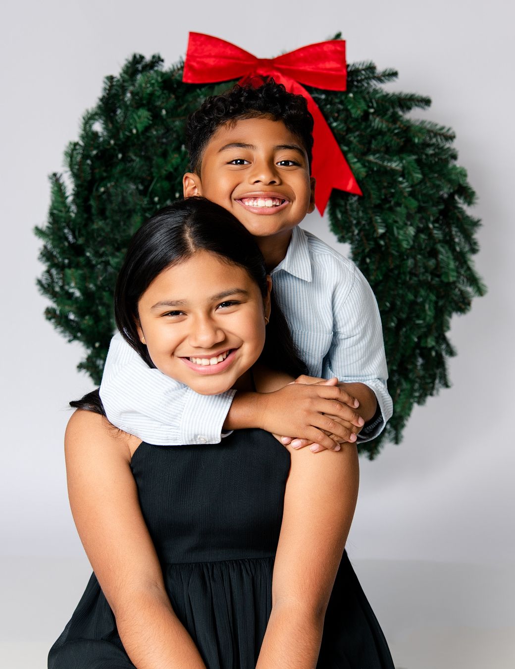 A child in a black dress poses playfully against a white background with a festive Christmas wreath.