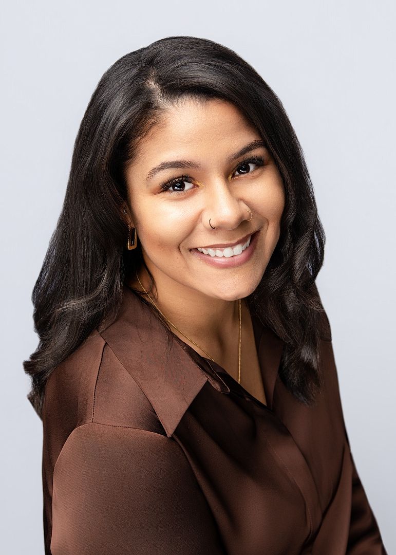 Professional headshot of a young woman with long black hair, wearing a brown satin blazer, smiling confidently.