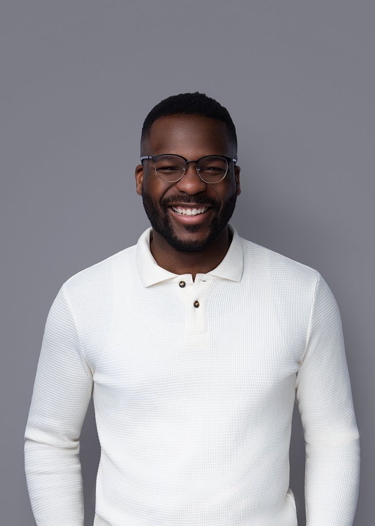 Smiling Black man wearing glasses and a white polo sweater poses against a gray background.