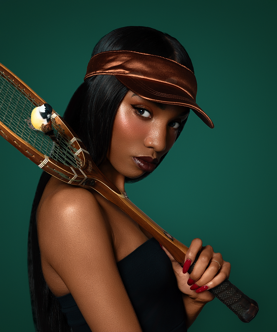 Stylish woman in bronze visor holding a wooden tennis racket against a teal background.
