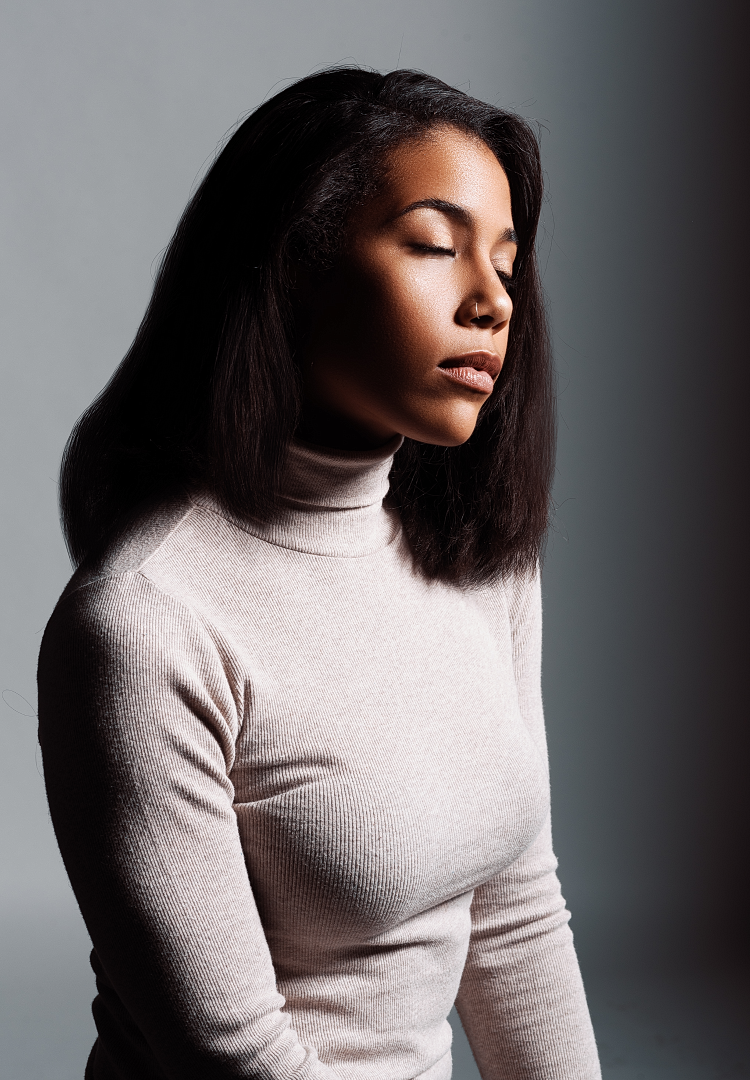Young woman with dark hair wearing a beige turtleneck sweater, eyes closed, posed against a gray background.