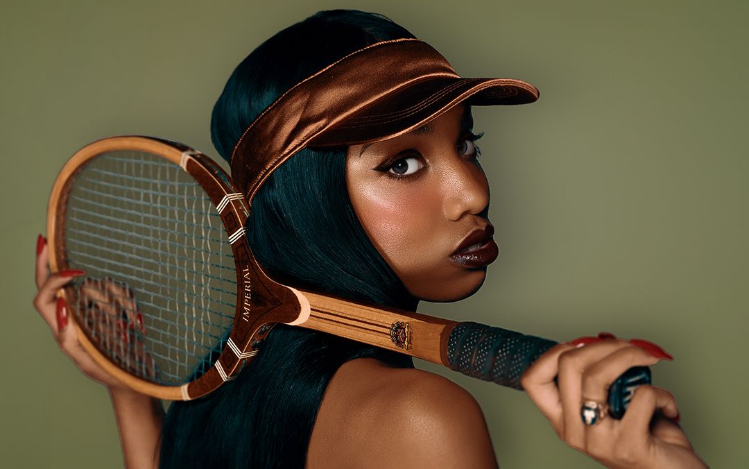 Young woman in bronze visor holding vintage wooden tennis racket, posing against olive green background.