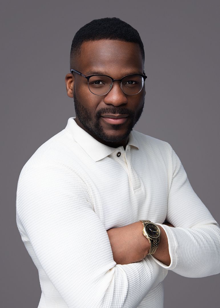 Professional man in white polo shirt with glasses and watch, arms crossed, posing against gray background.