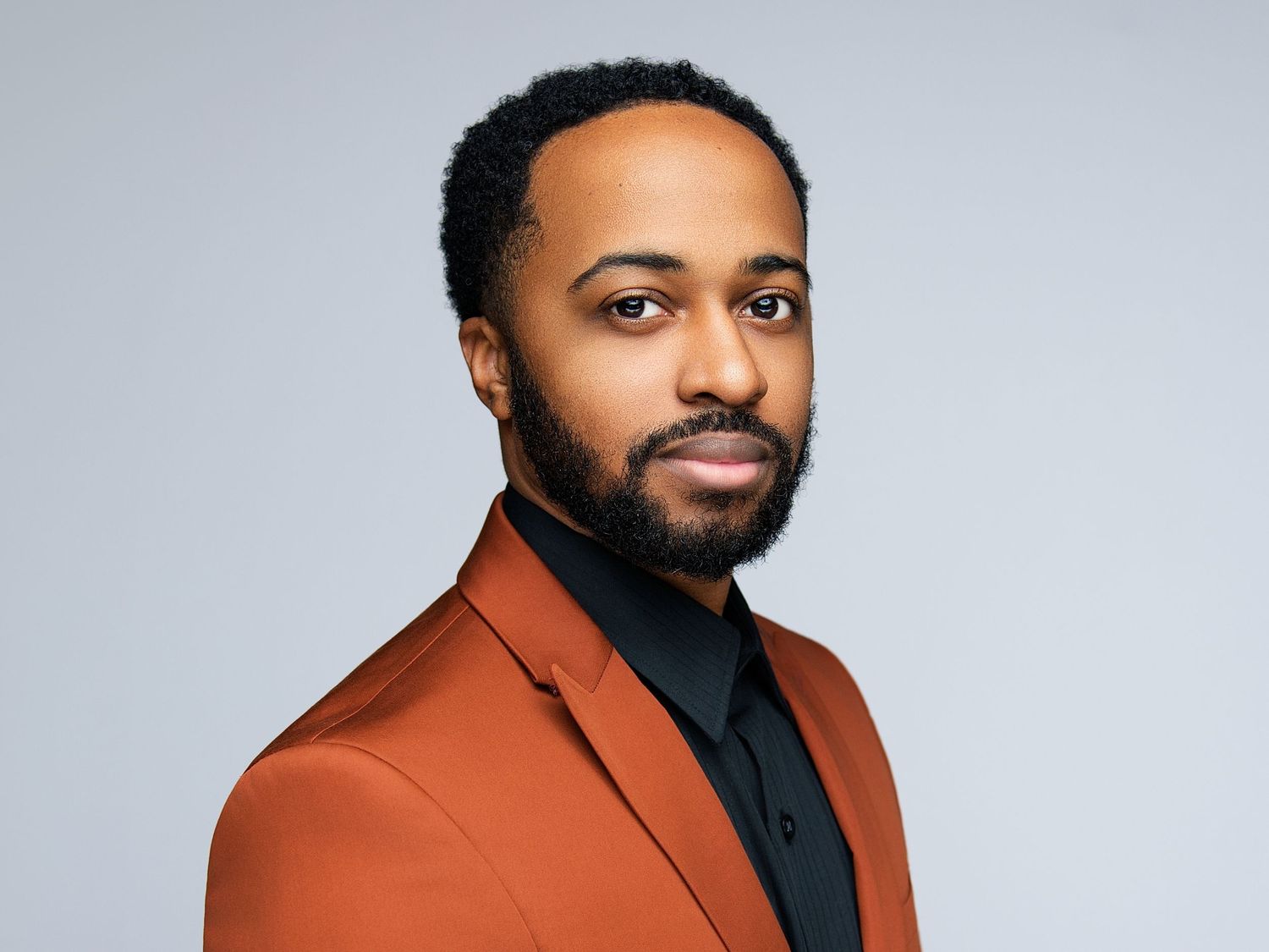 Professional headshot of person in orange blazer and black shirt against light gray background.
