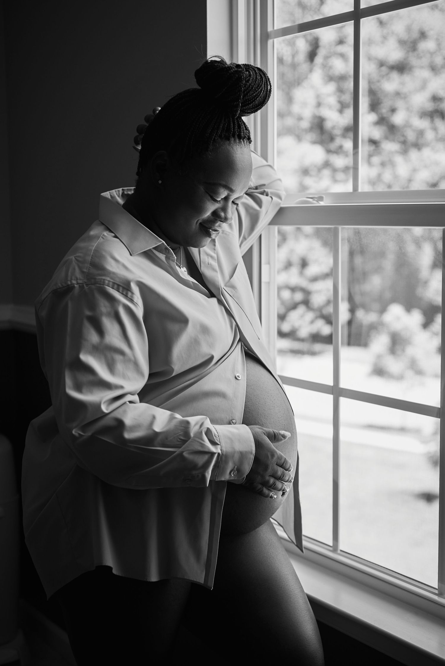 Black and white maternity photos by window with dramatic natural lighting.