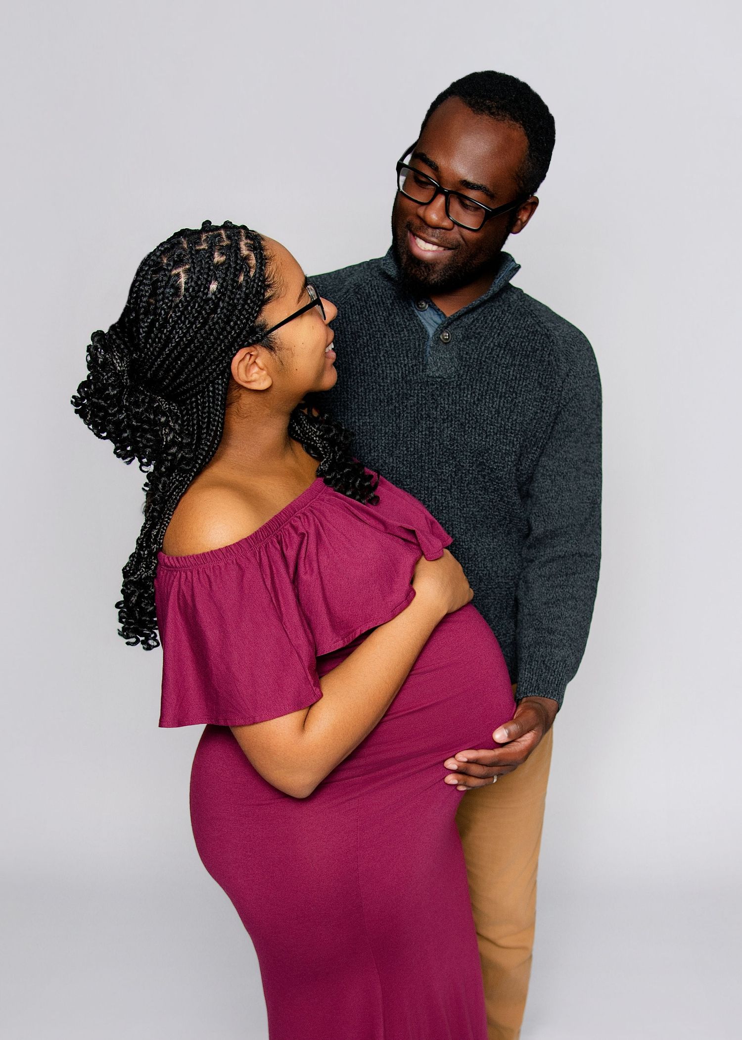 A smiling couple shares a tender maternity moment, with the man embracing the pregnant woman in a magenta off-shoulder dress.