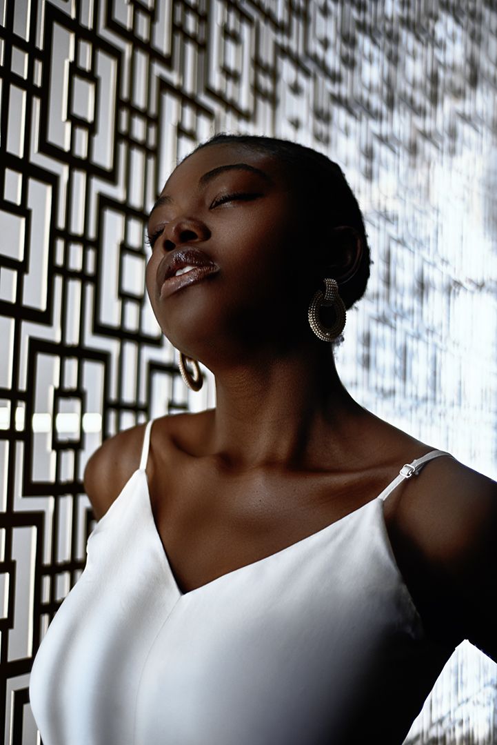 A confident Black woman in a white camisole poses with eyes closed, wearing hoop earrings against a geometric patterned wall.