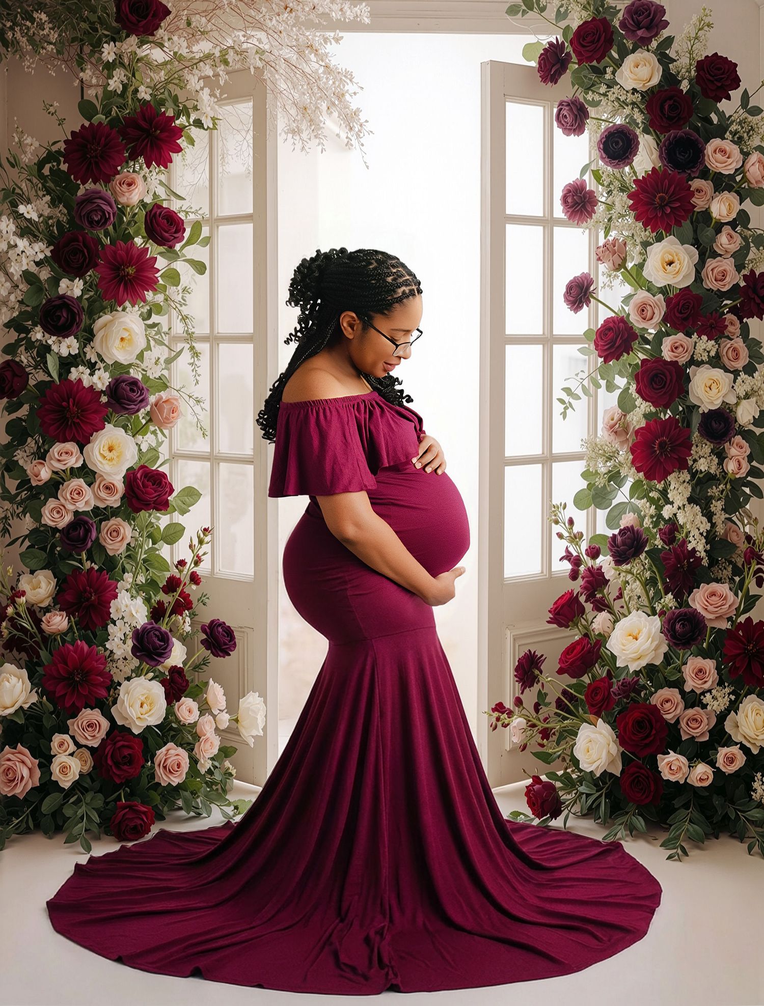 Pregnant woman in burgundy off-shoulder gown poses gracefully among lush floral arrangements of roses in a bright studio.