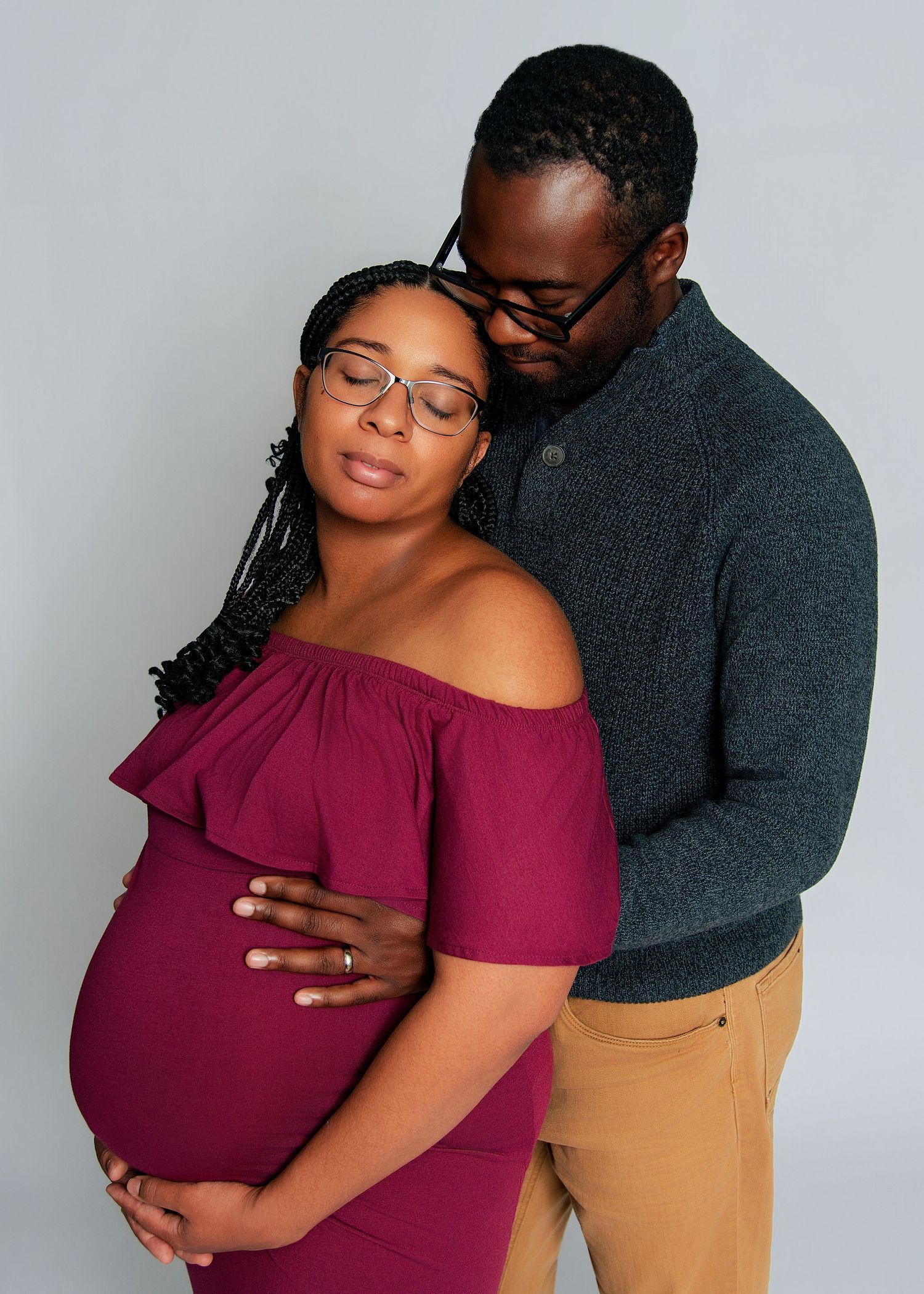 A couple shares a tender maternity photo, the pregnant woman in a burgundy off-shoulder dress, her partner embracing her from behind.