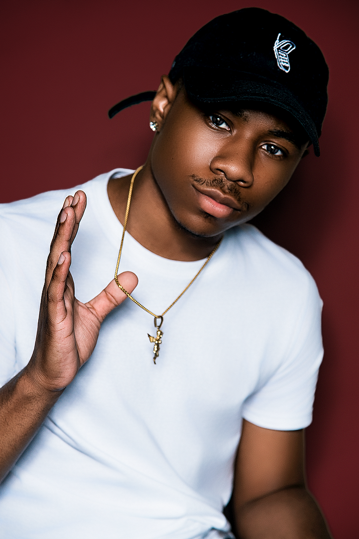 Young man in white tee and black cap wearing gold chain necklace poses against dark red background.