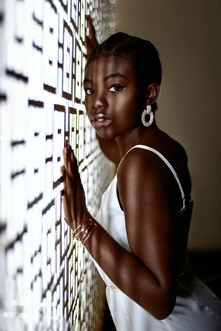 Young Black woman in white dress and hoop earrings leaning against a decorative wall, looking at camera.