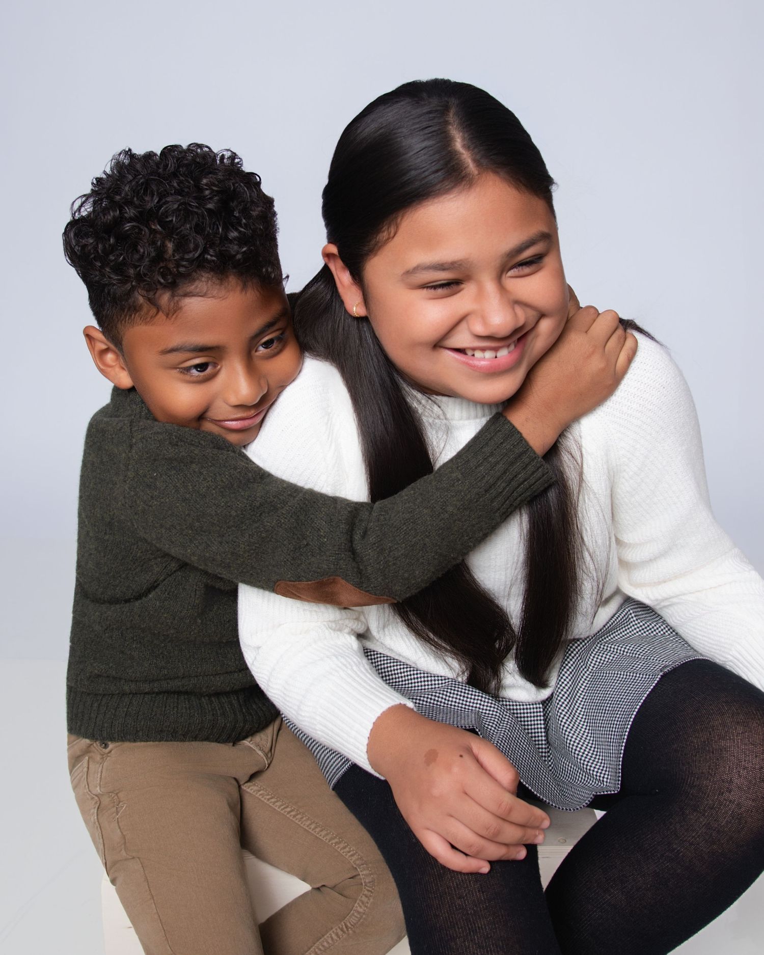 Two children hugging warmly in a studio, a boy in olive sweater and a girl in white sweater, smiling together.