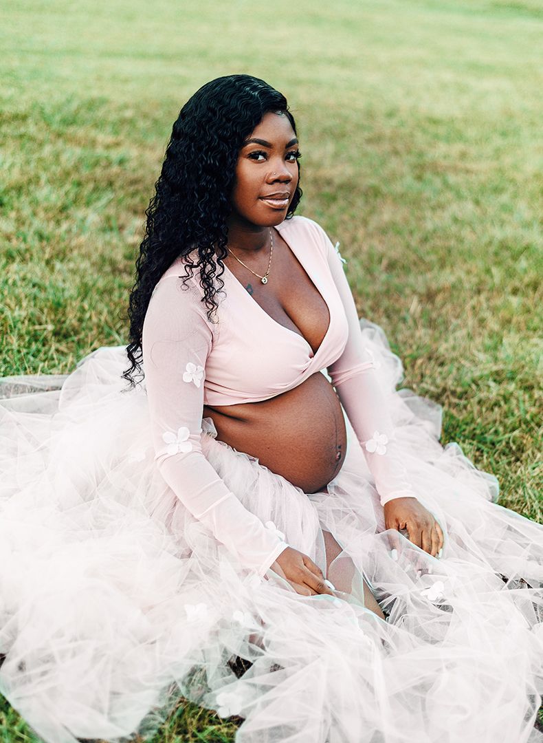 Maternity portraits in pink wrap top and white tulle skirt on grassy field.