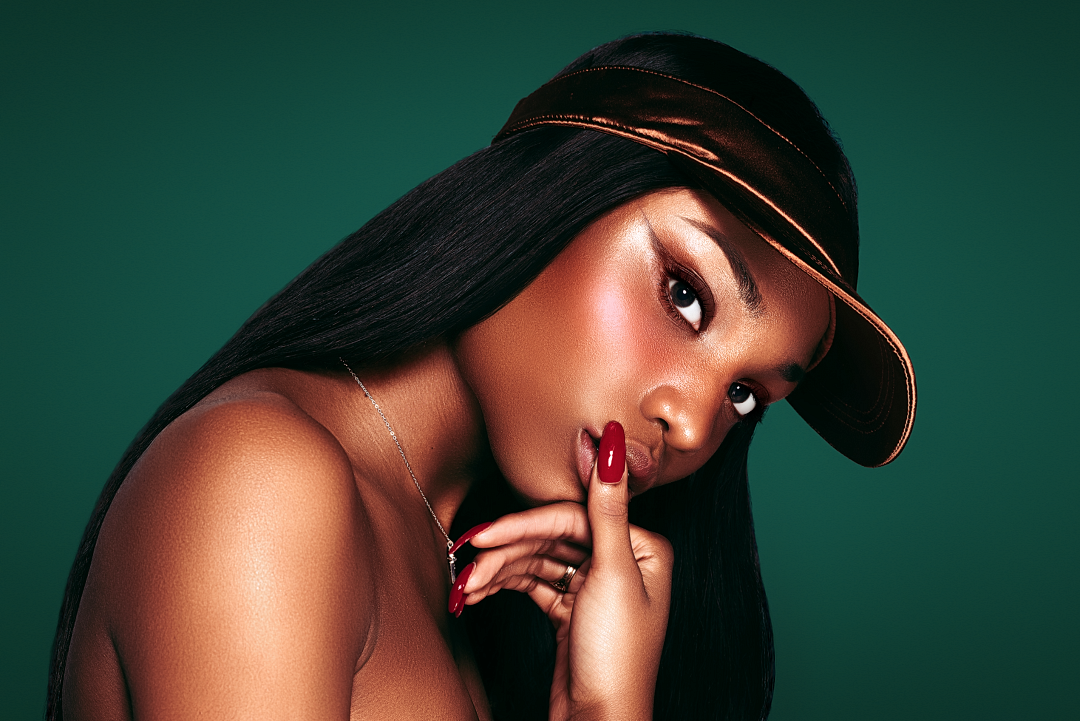 Glamorous Black woman with sleek hair, gold visor cap, red nails, posing against teal background.