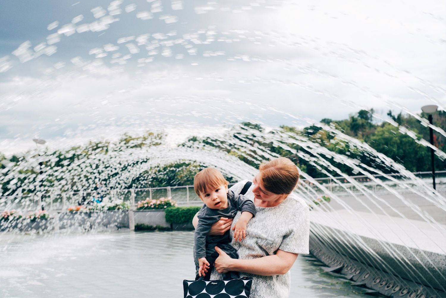 A father holds his toddler near a splashing fountain outdoors under a partly cloudy sky.