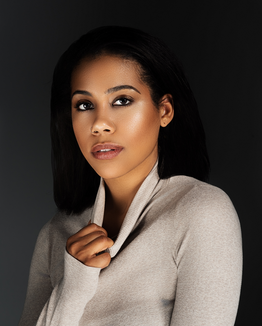 Young Black woman in beige turtleneck sweater poses confidently against dark background with sleek straight hair.