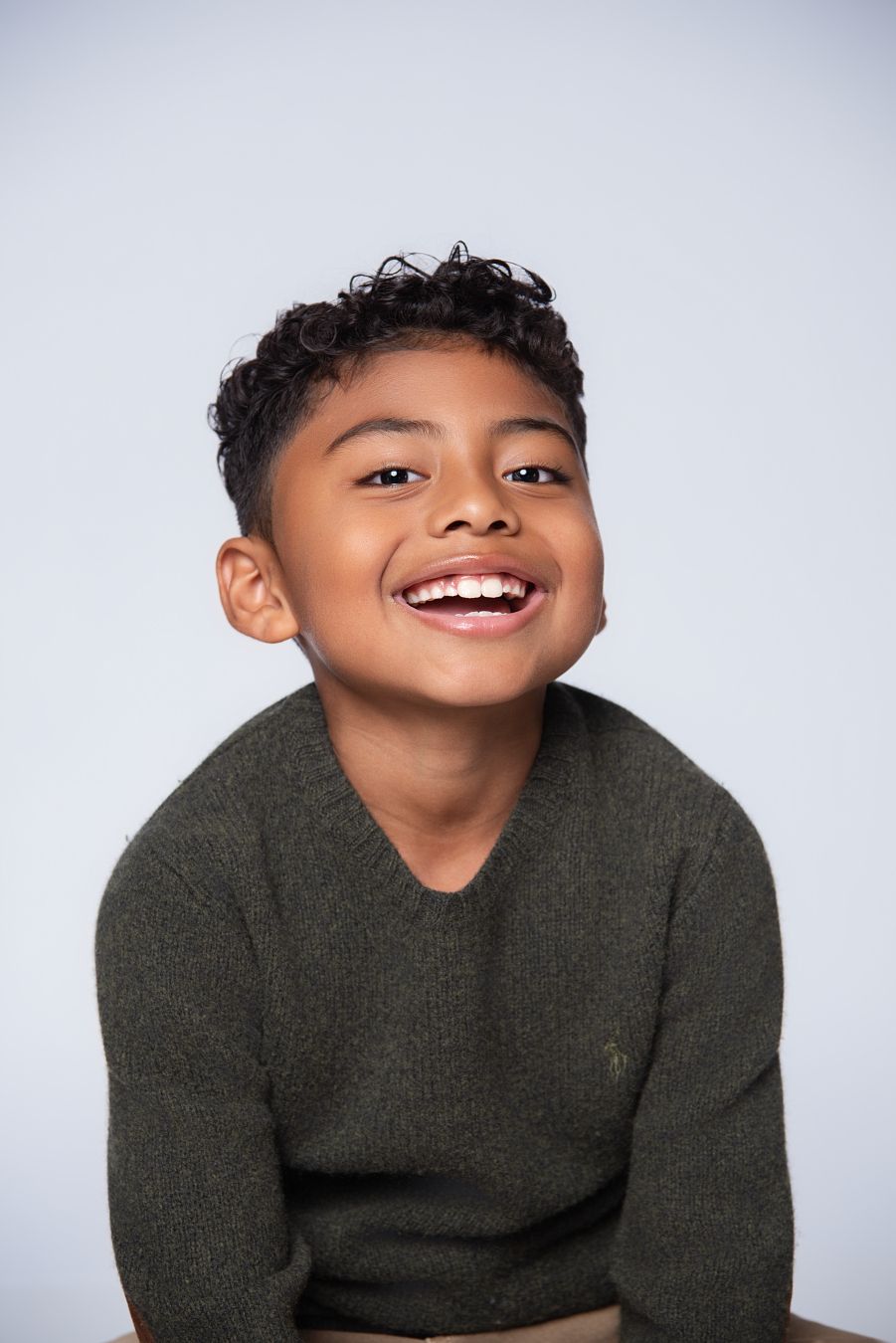 A child in a dark green sweater smiles brightly against a light gray background.