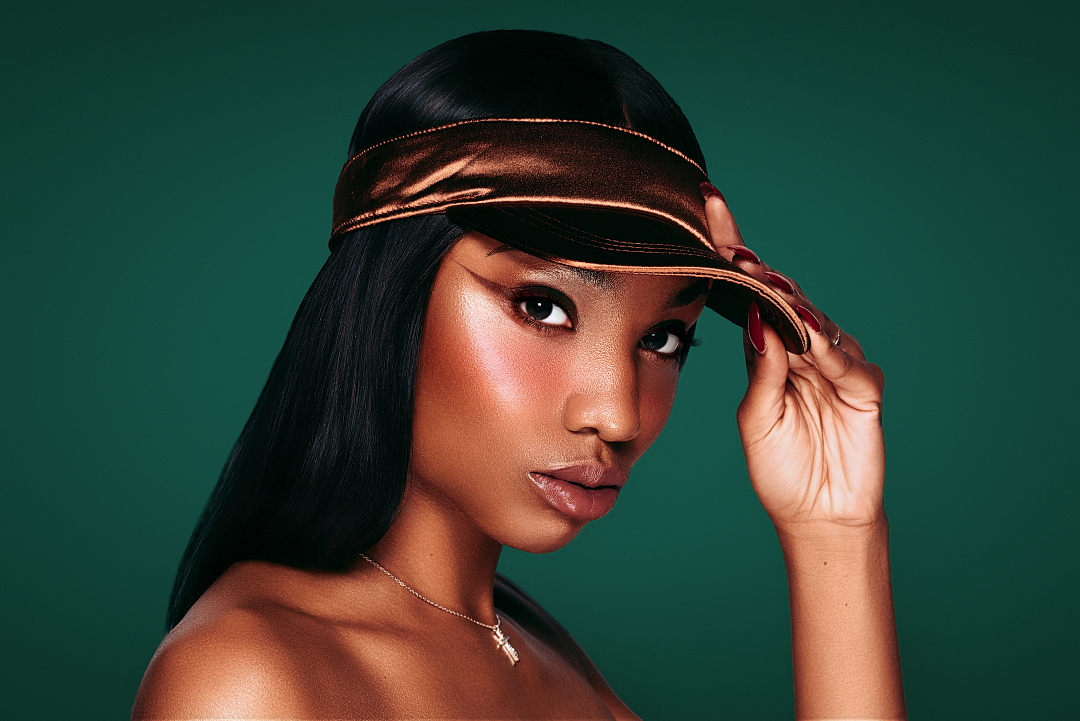 Young woman with glowing skin wearing a bronze metallic visor against a teal background.