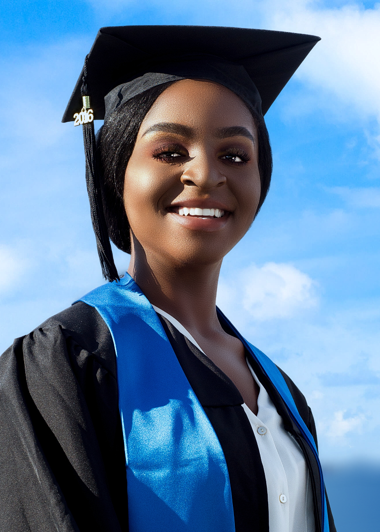 Young woman in black graduation gown and cap with blue sash smiling against a blue sky backdrop.