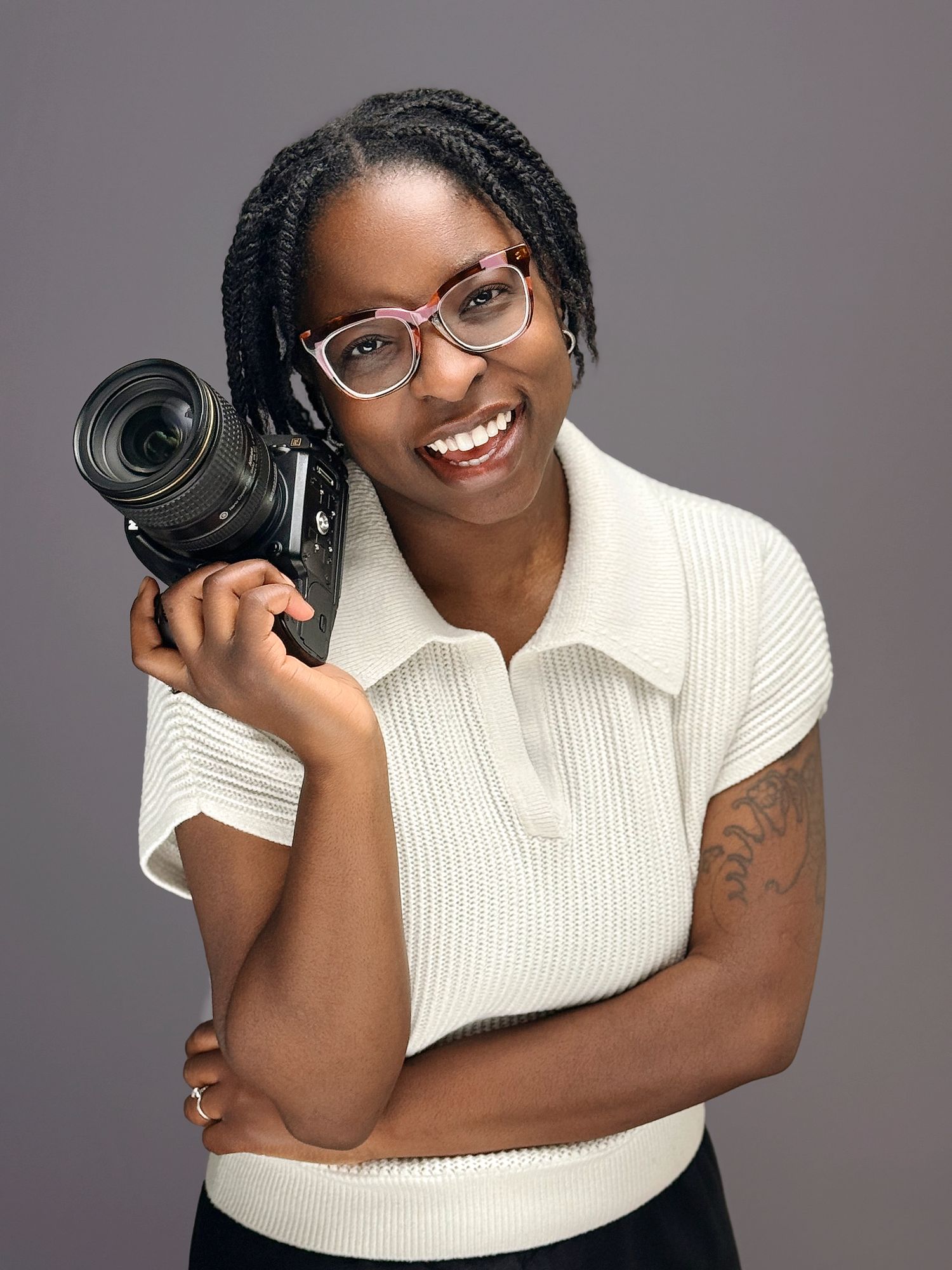 Professional photographer in a white collared top holds a camera lens while posing against a gray studio background.