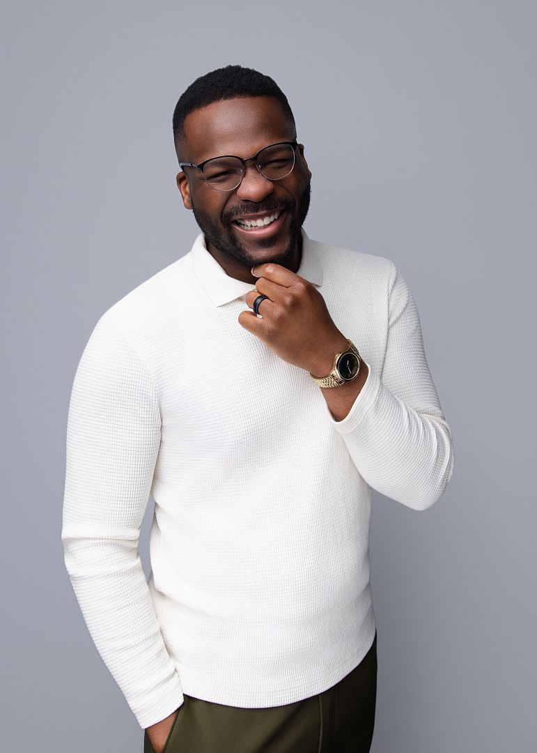 Smiling Black man in white ribbed sweater and glasses, posing confidently against a gray background.