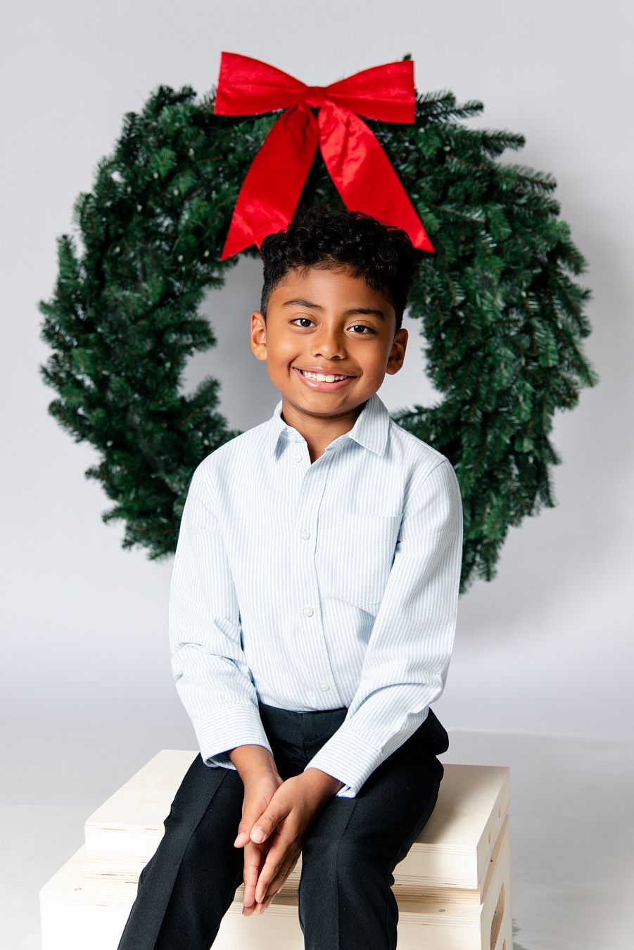 Holiday portrait series showing a young child in white shirt and dark pants seated with evergreen wreaths and red bows.