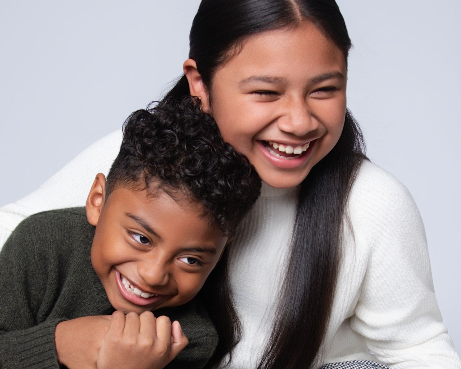 Two smiling children laughing together in a studio portrait, wearing casual clothes against a light gray background.