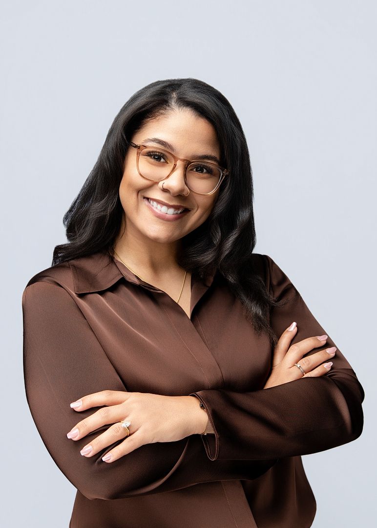 Smiling woman with glasses and brown satin blouse, arms crossed, posing confidently against a light gray background.