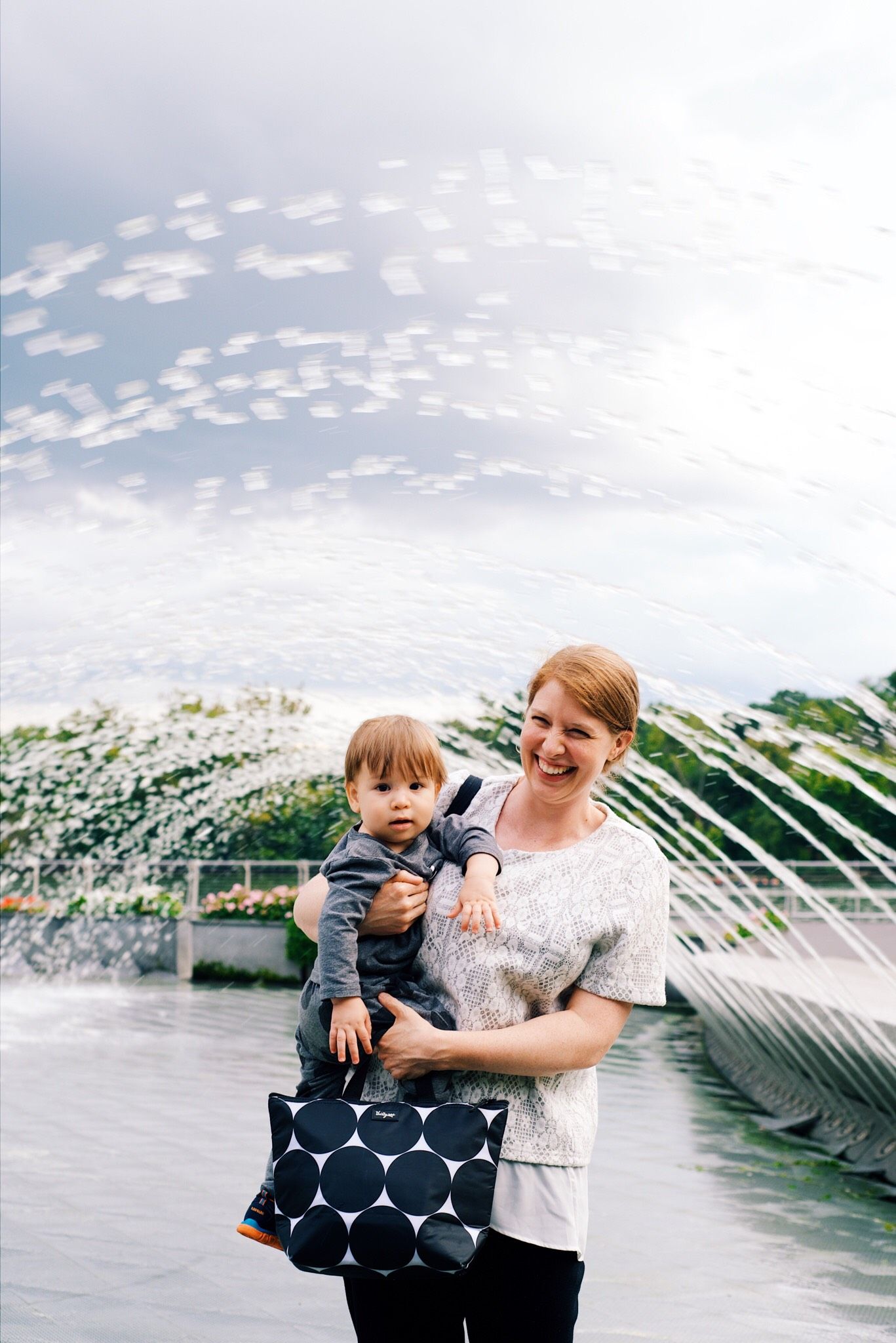 A smiling woman holds a baby outdoors near a fountain, carrying a navy polka dot tote bag on a cloudy day.