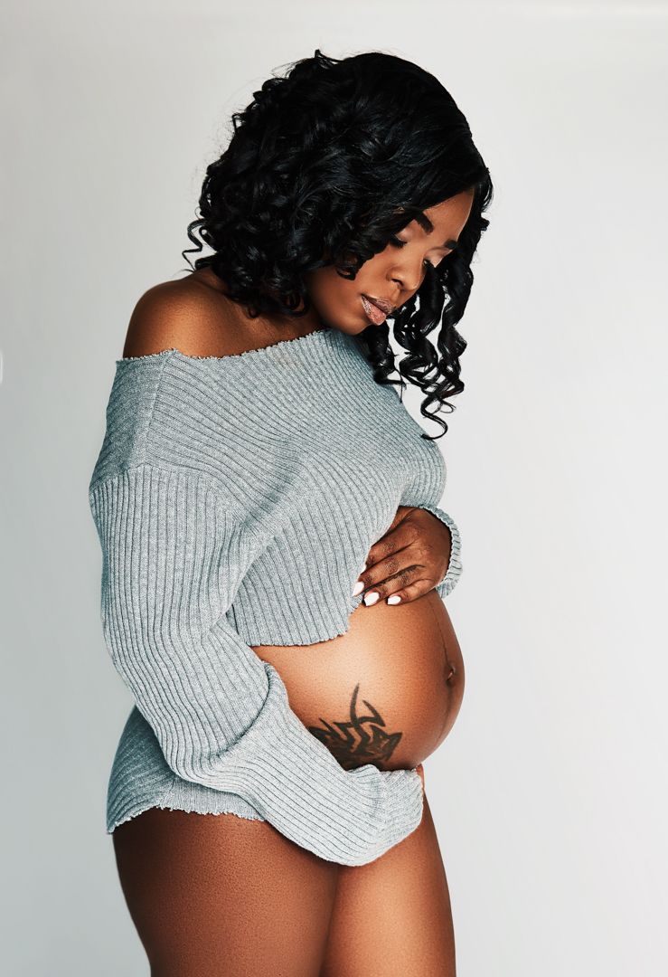 Maternity portrait showing pregnant woman in gray sweater against white background.
