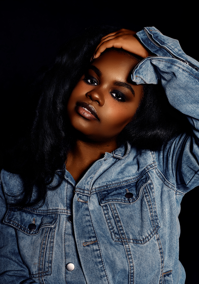 A young Black woman in a denim jacket poses confidently against a dark background with smoky eye makeup.