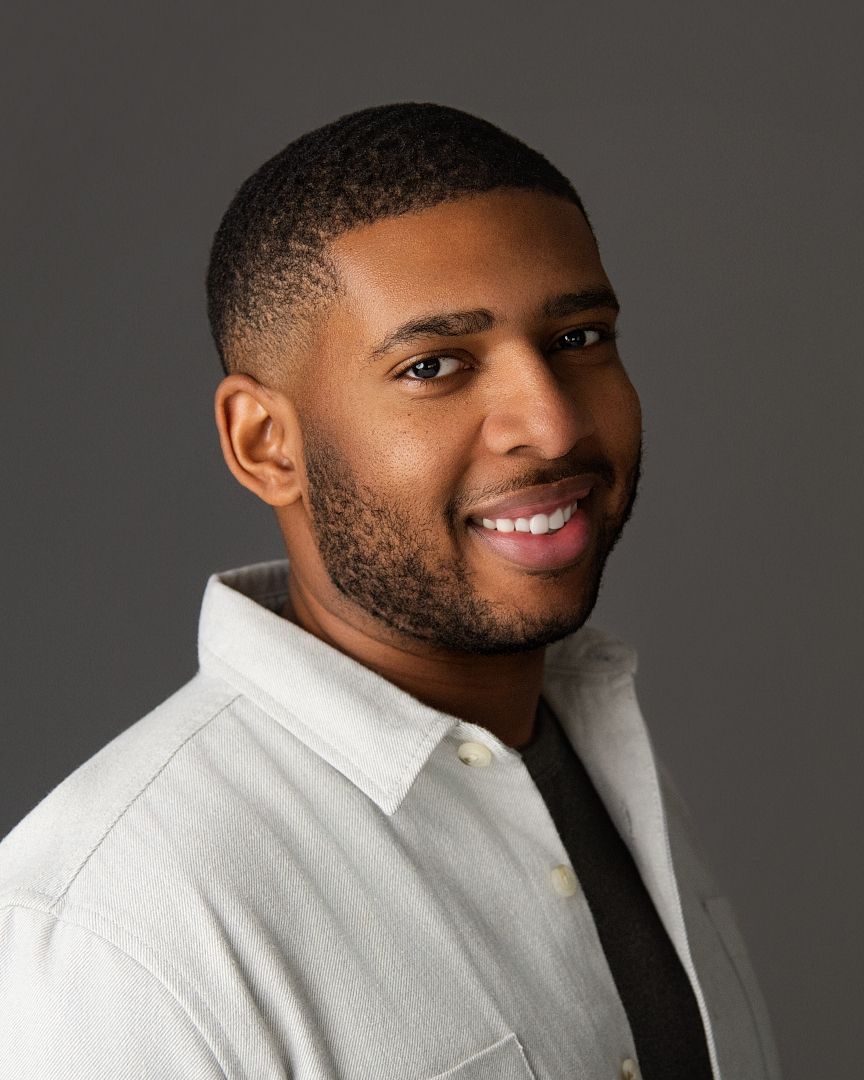 Professional headshot of a person wearing a white collared shirt against a dark gray background, smiling warmly.