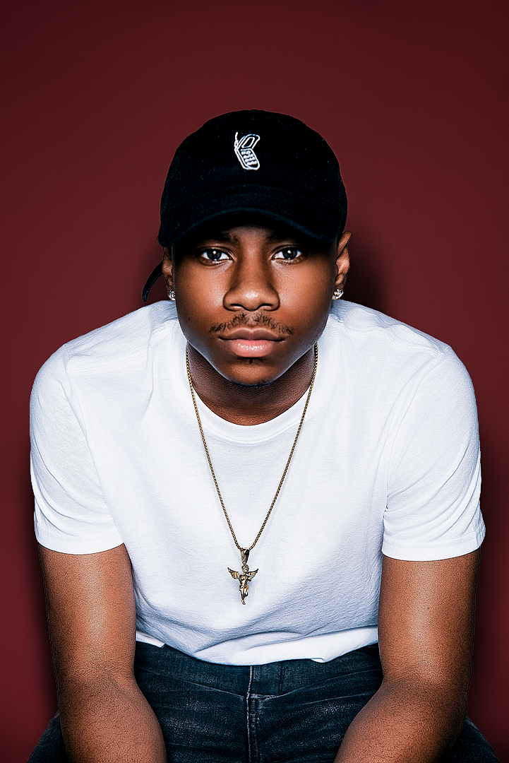 Young man in white t-shirt and black baseball cap with gold necklace sits against dark red backdrop.