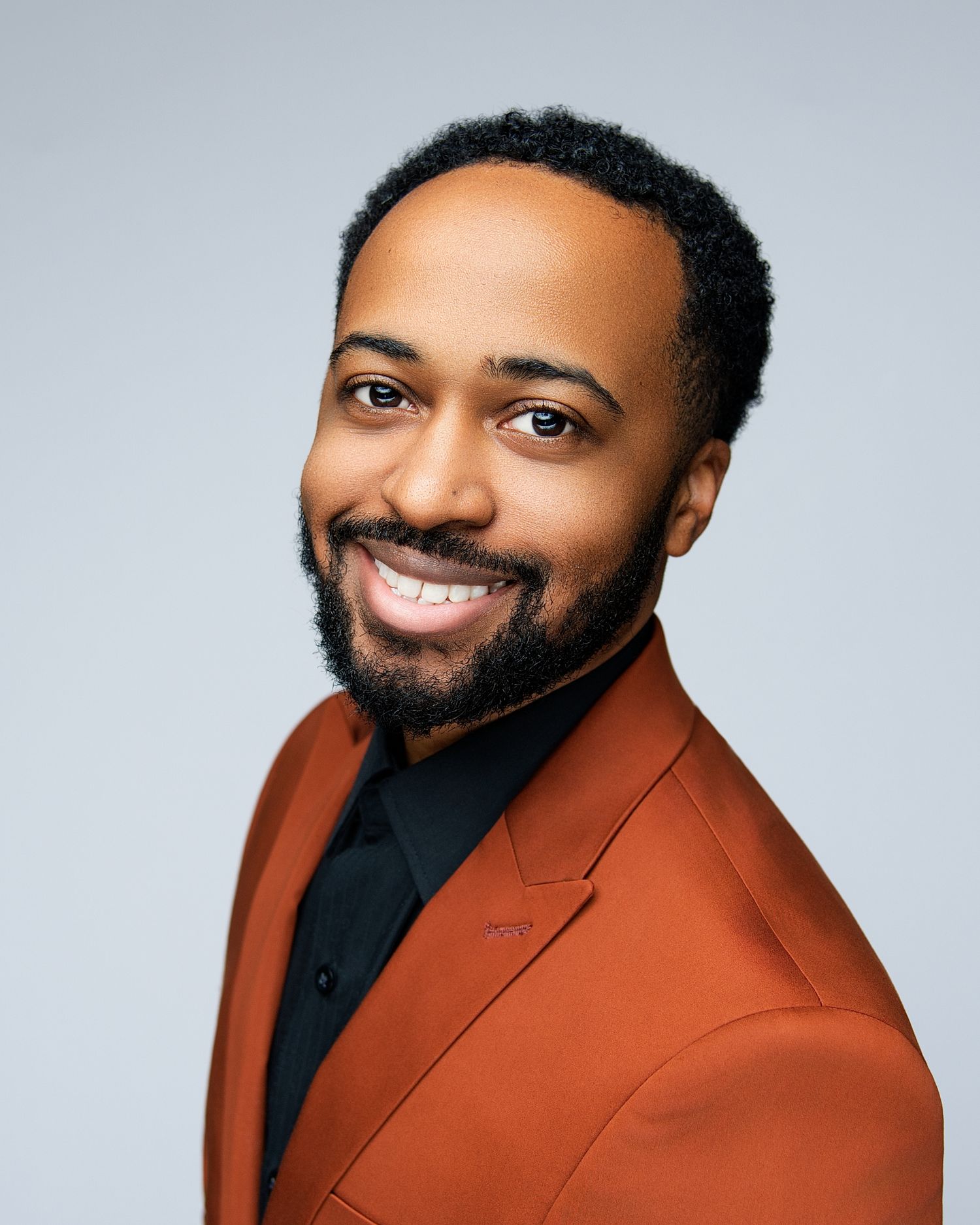 A man wearing a rust-colored blazer and black shirt smiles warmly in a professional headshot series.