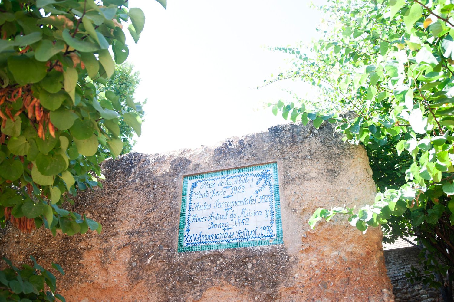 A weathered terracotta wall with a decorative blue tile plaque mounted on it, framed by green foliage.