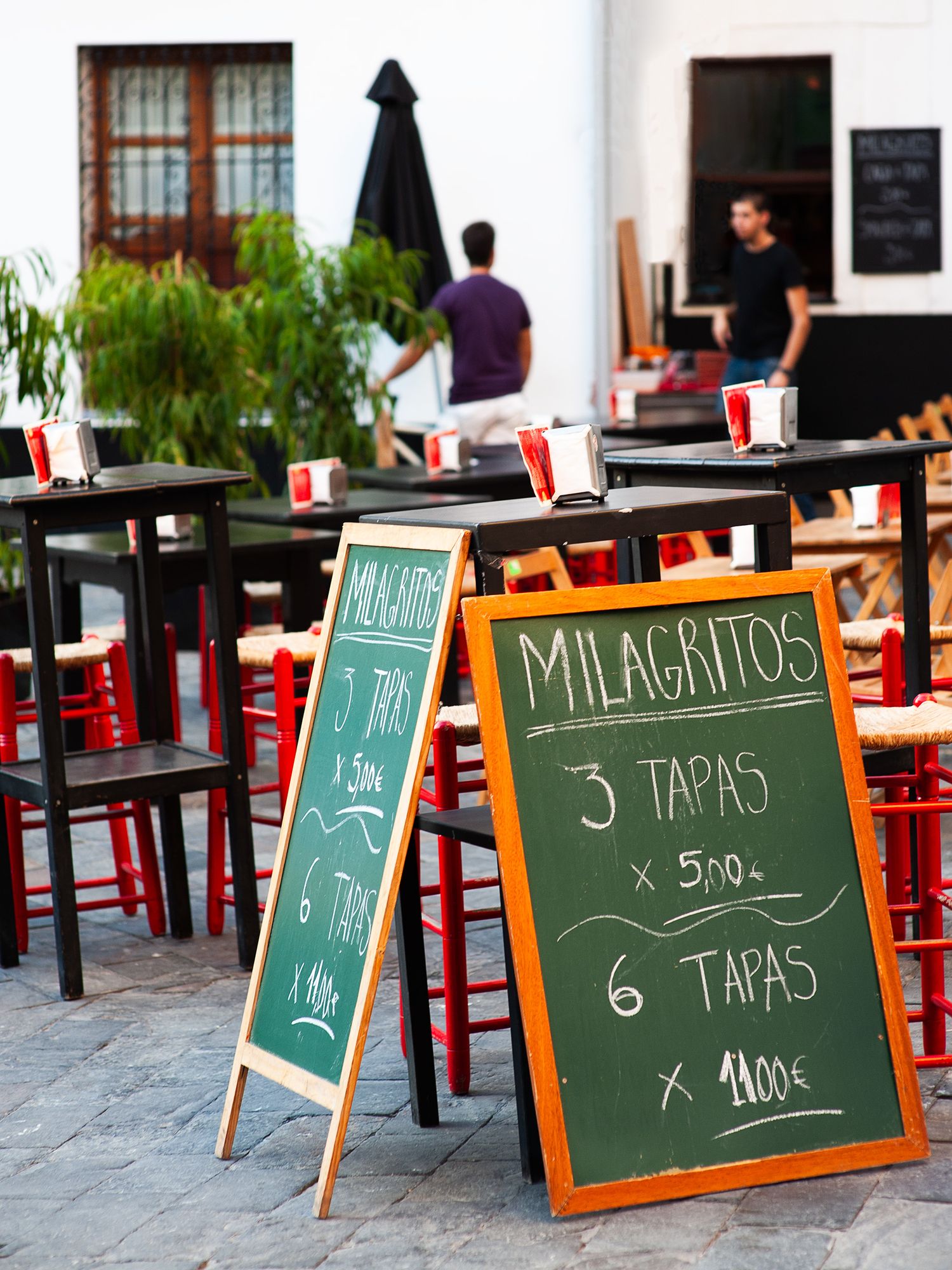Outdoor cafe seating area with wooden tables and chalkboard signs displaying menu items for tapas and milagros.