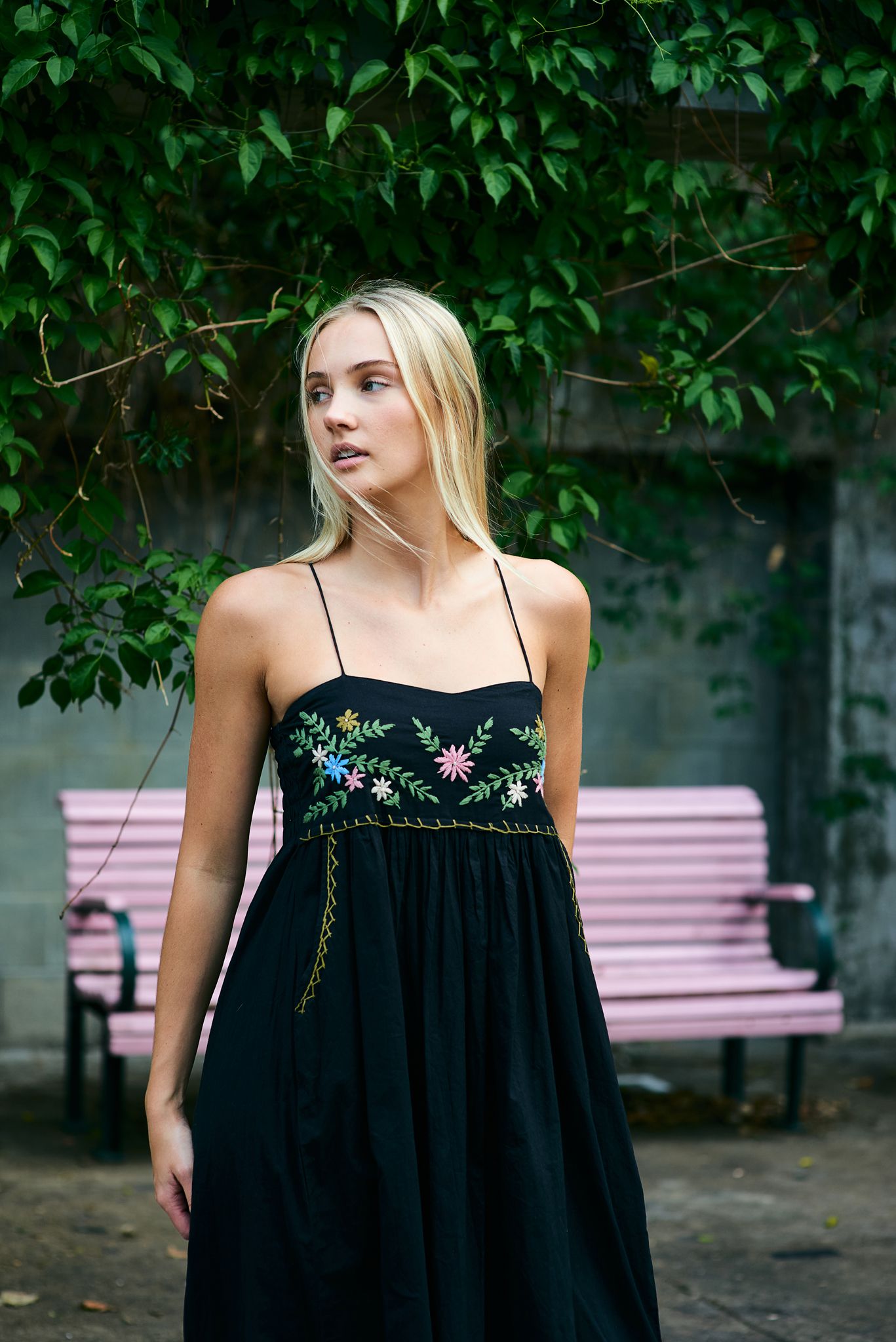 Woman in black sundress with floral embroidery poses outdoors against leafy green garden backdrop and pink bench.