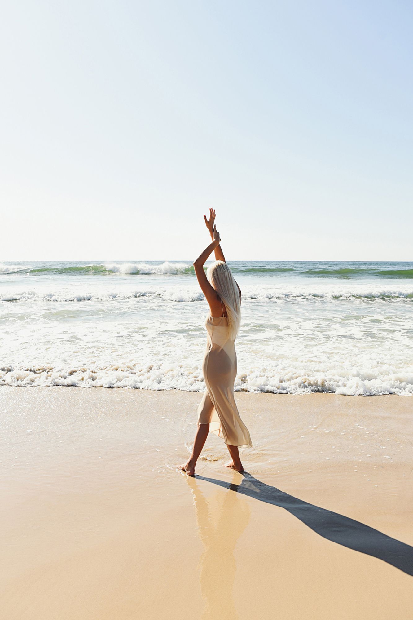 Person in light clothing dances joyfully on sandy beach with ocean waves in background.
