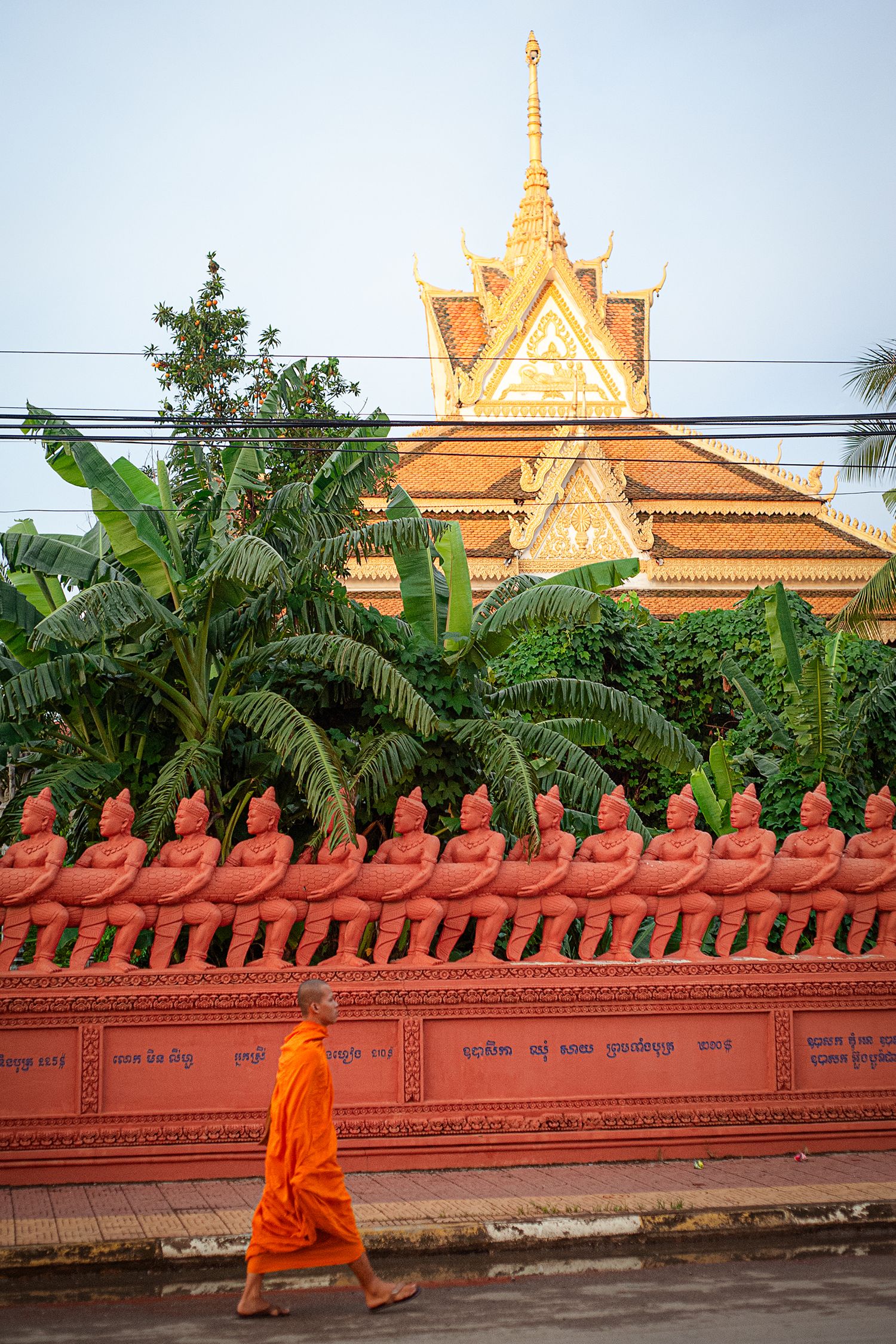 Buddhist monk in orange robes walks past ornate temple wall with decorative red statues in Phnom Penh, Cambodia.