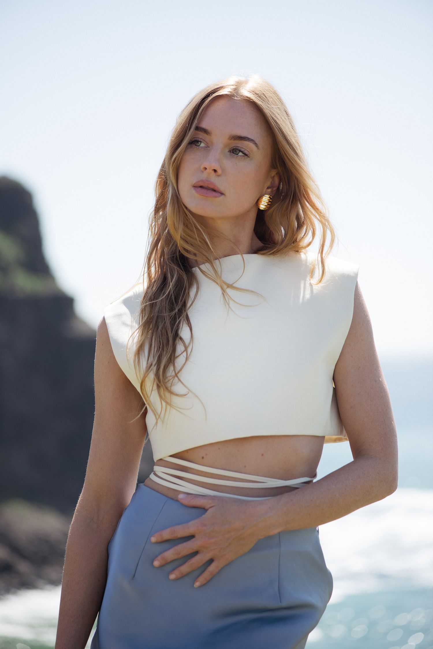 Fashion model in white crop top and gray pants poses against coastal cliffs and ocean backdrop.
