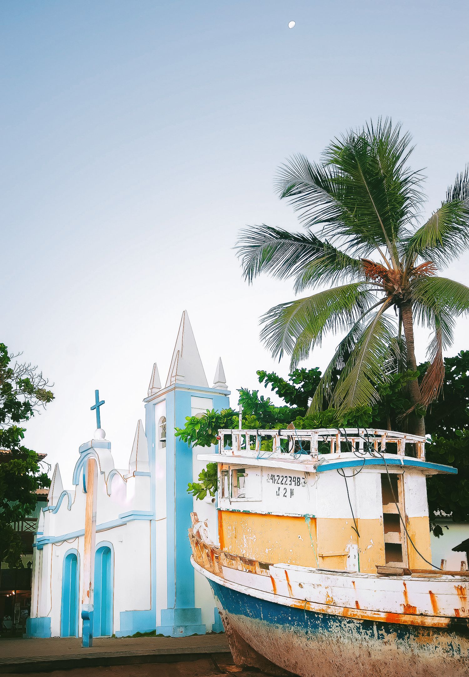 A coastal church with blue and white walls, palm trees, and an old wooden boat on the beach.