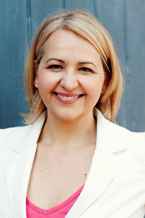 Professional portrait of someone in white blazer and pink top smiling against blue background.