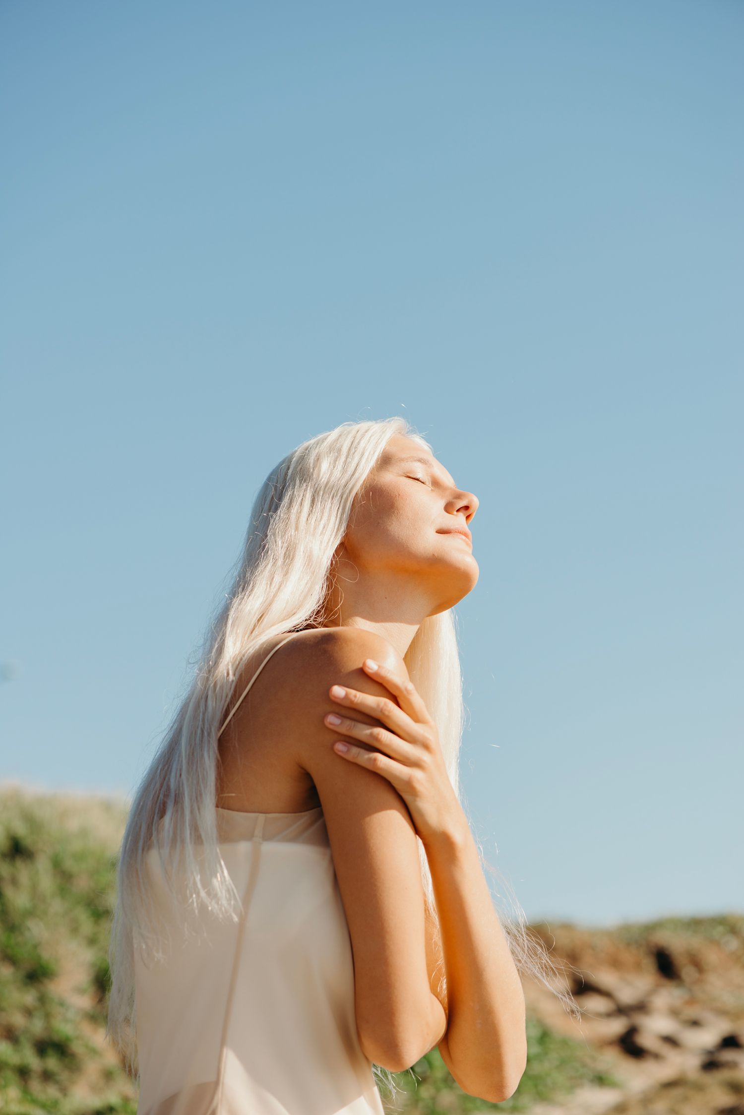 Person with platinum blonde hair poses against blue sky in white clothing while touching their neck in sunlight.